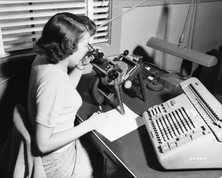 A human computer at work at NASA Langley Research Center in 1952, using a microscope to read data from film while a Friden calculating machine sits beside her.